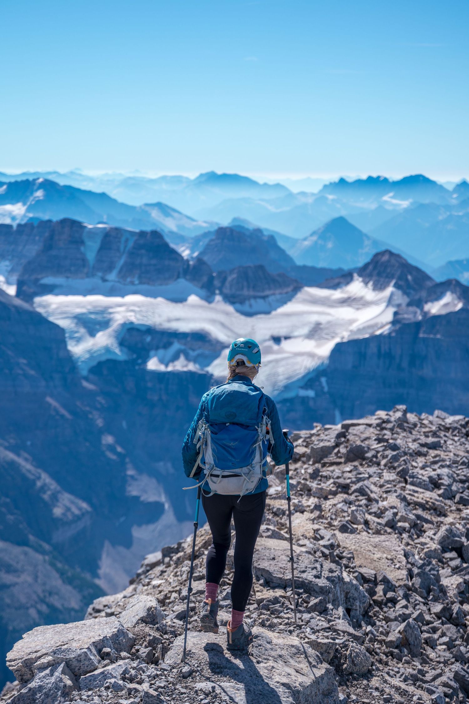 Hiker standing on rocky summit overlooking snow-capped mountain peaks stretching into the distance.