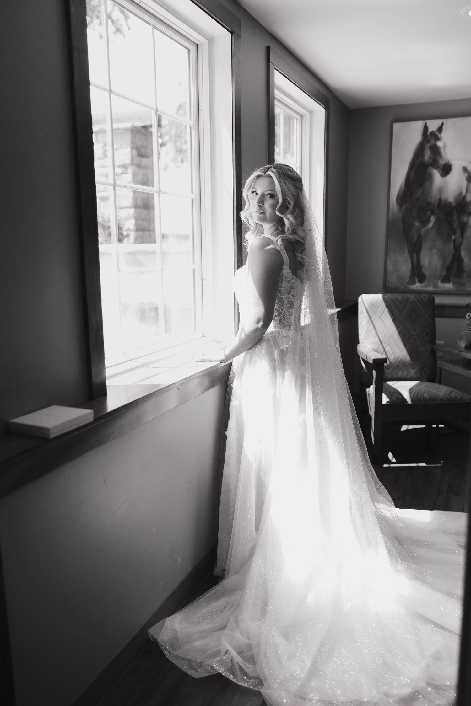 Black and white series showing a bride in a flowing white wedding gown near large windows in an interior space.