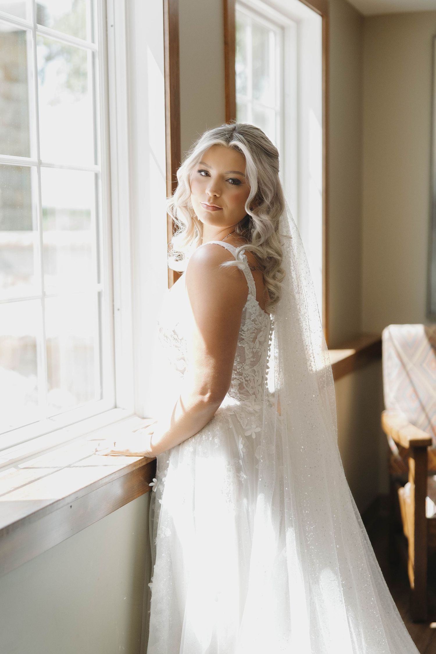 Bride in elegant white lace wedding dress and veil gazes out window in soft natural light.