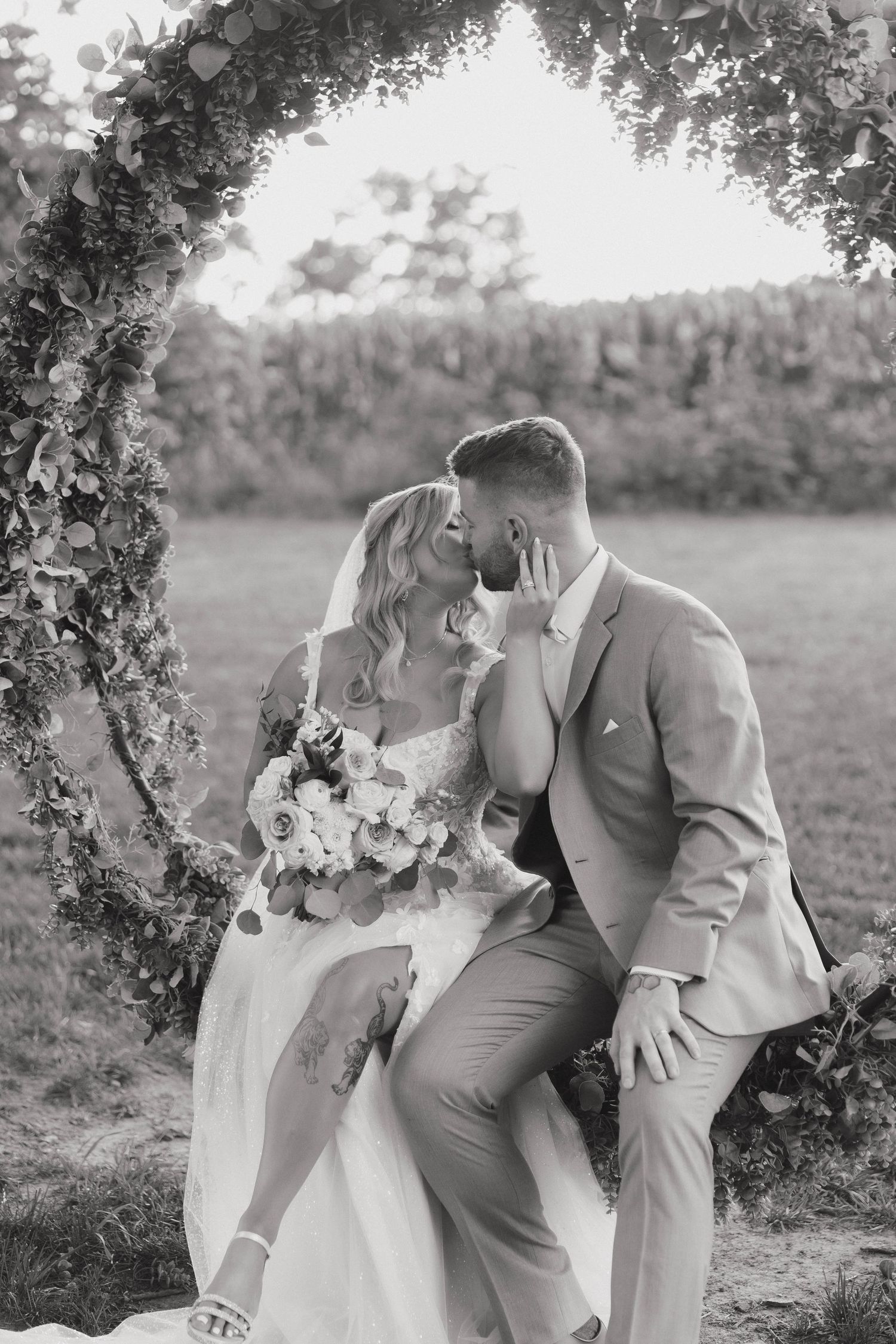 A romantic black and white wedding photo of a couple sharing an intimate moment under a floral arch in a garden setting.