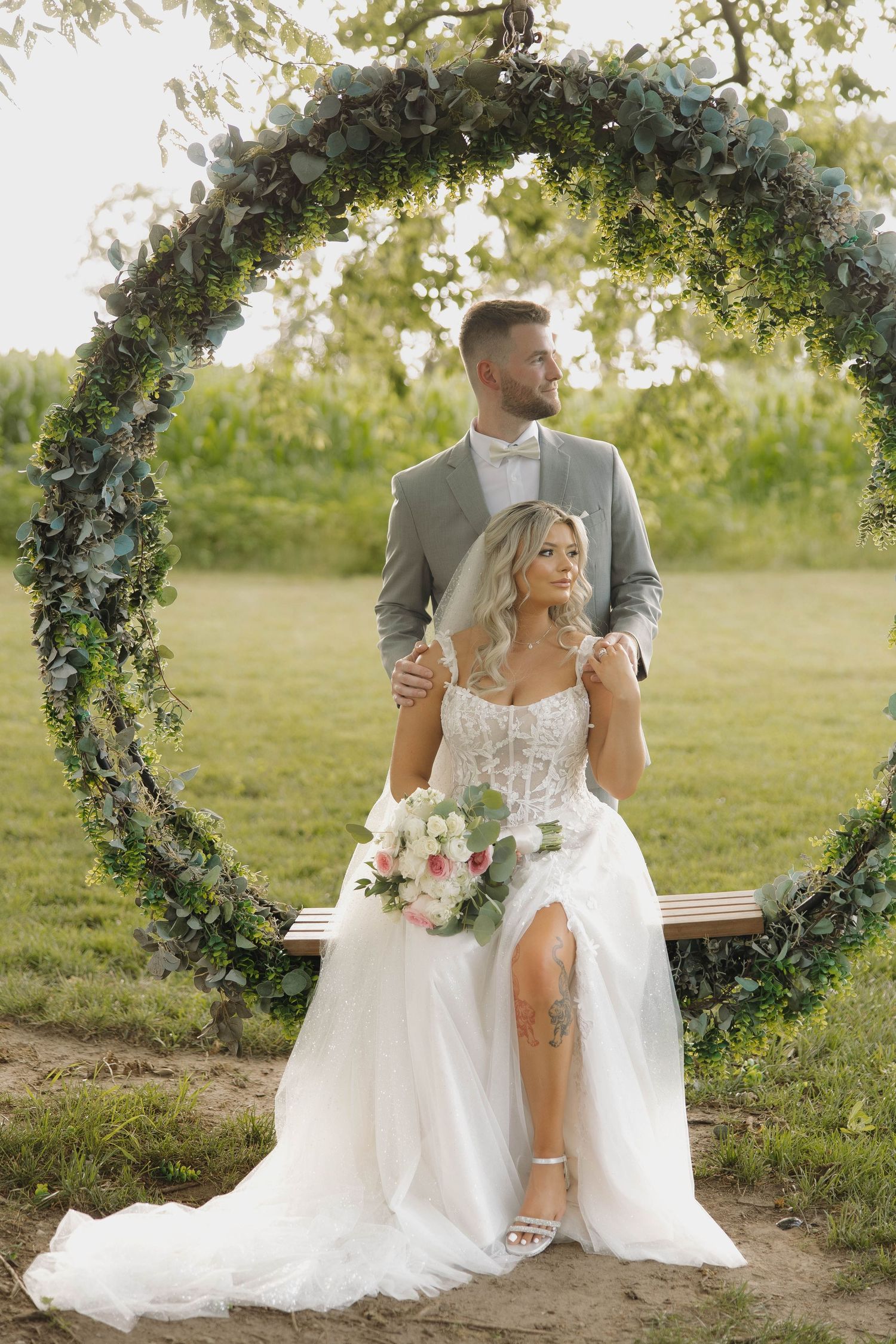 Newlywed couple sits together under floral arch in romantic outdoor garden wedding setting during golden hour.
