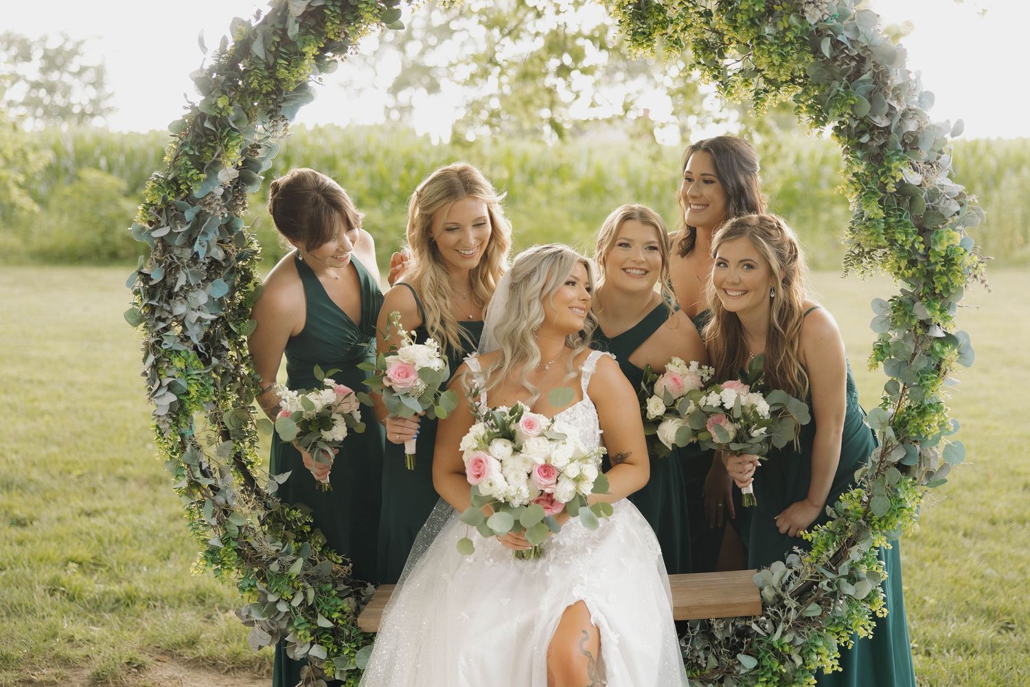 A wedding party poses outdoors under a circular floral arch in a sunlit field during golden hour.