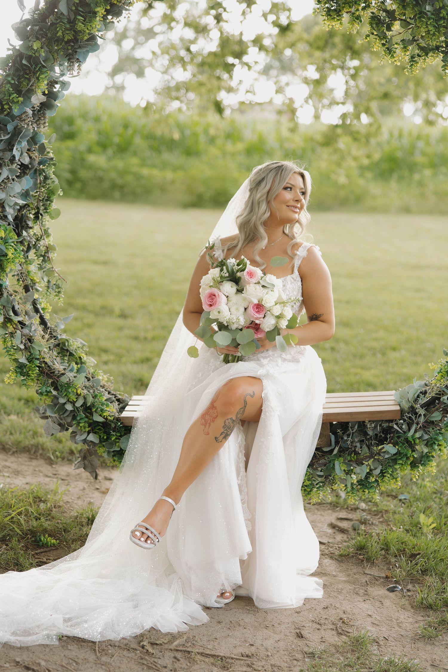A bride in a white wedding gown poses gracefully on a wooden bench in a natural outdoor setting.