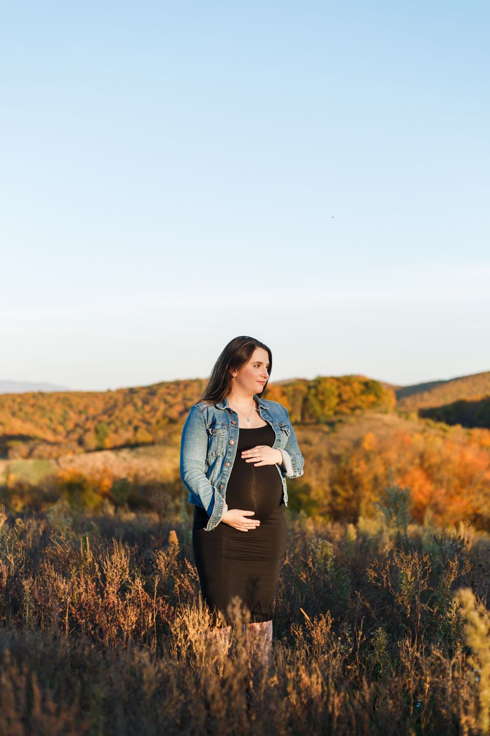 A woman in denim jacket stands in a golden autumn field with mountains during sunset.