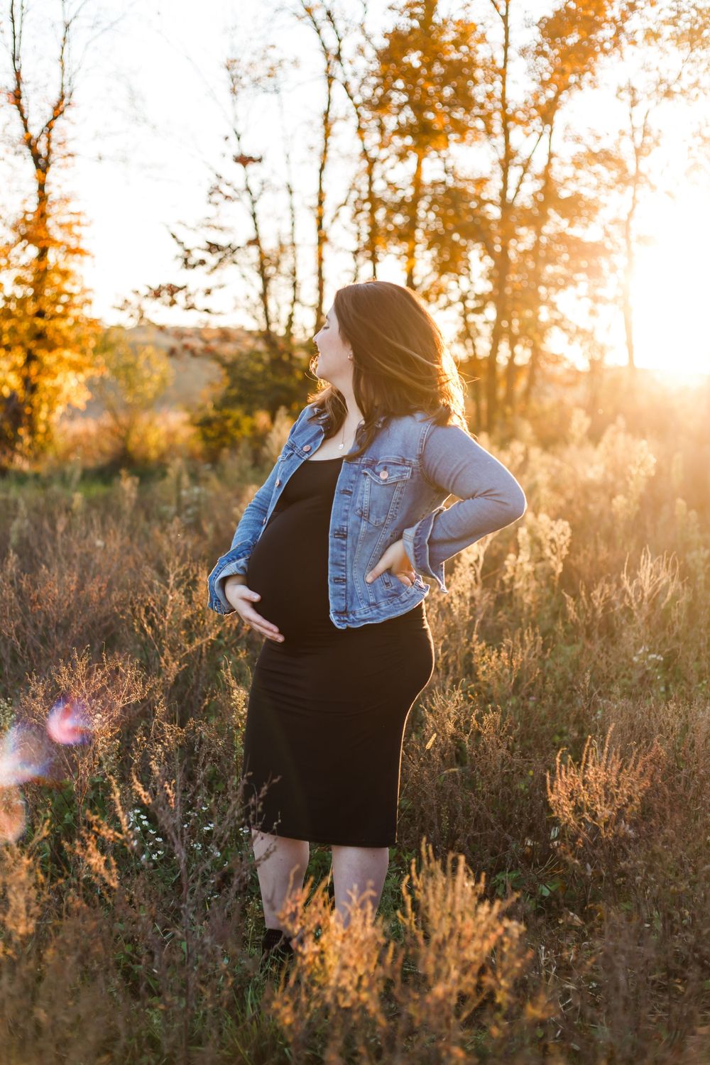 Silhouette of pregnant woman in black dress and denim jacket standing in sunlit meadow at sunset.