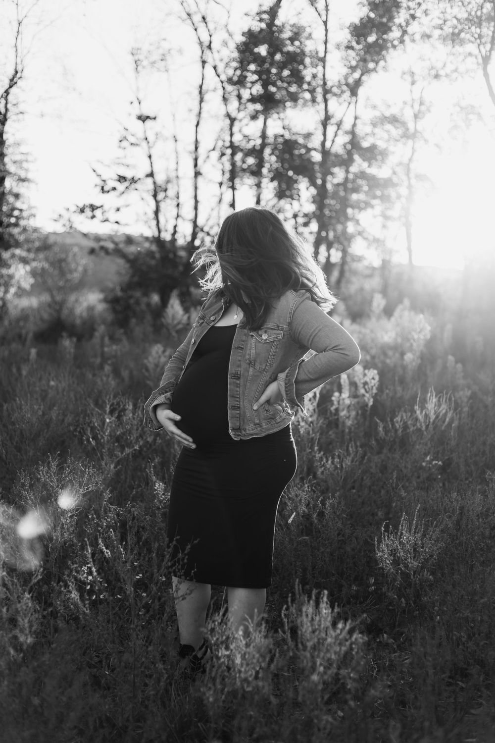 Artistic black and white photo of a woman in a dark dress walking through tall grass at sunset in a field.