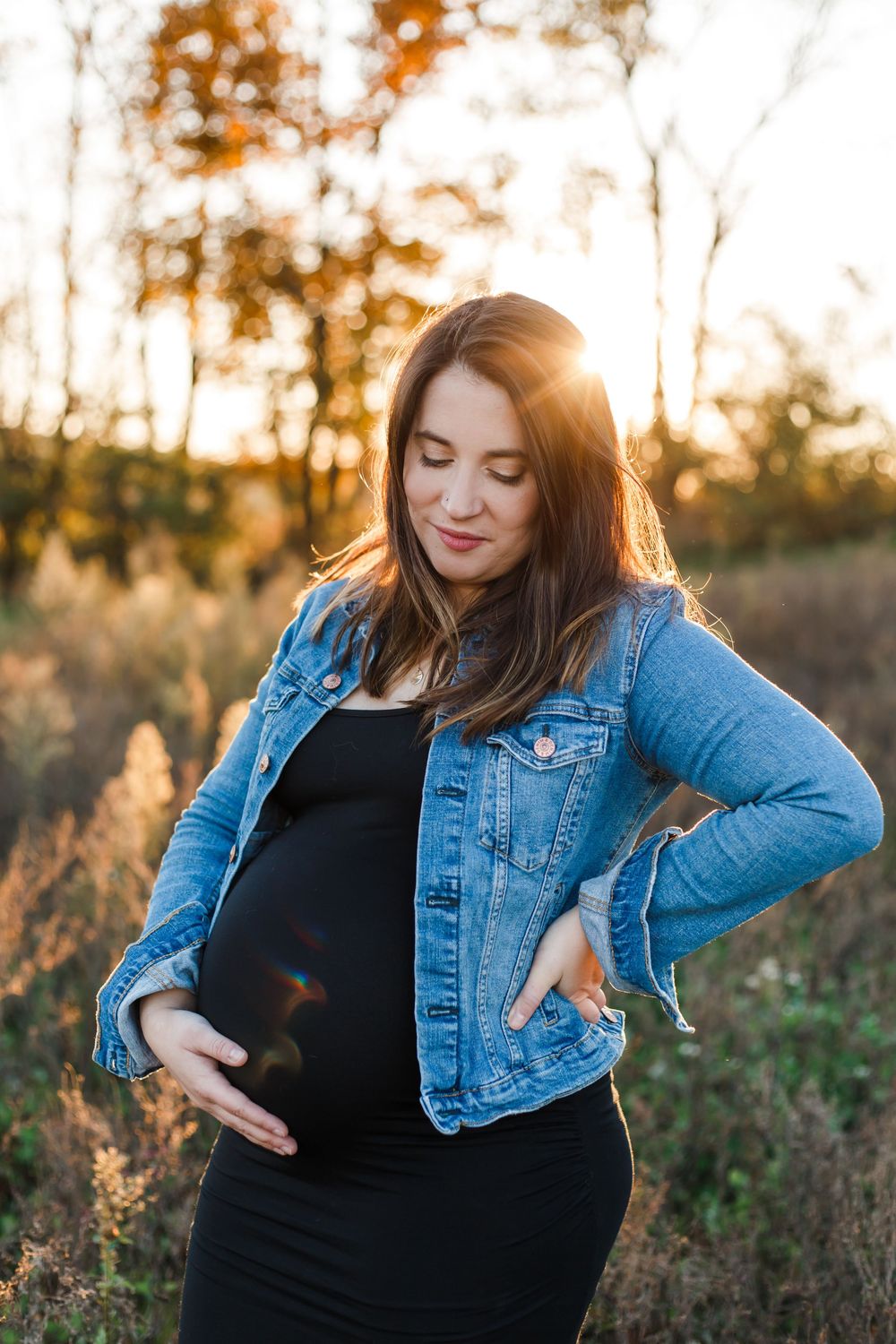 Expectant mother in denim jacket poses in a sunlit field during golden hour.