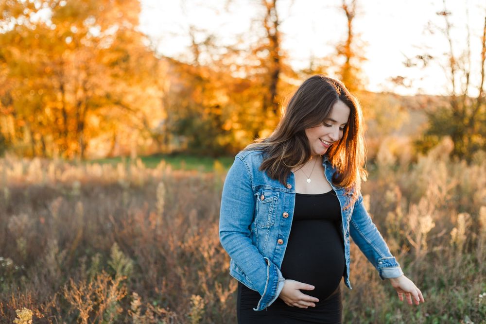 Expectant mother in denim jacket stands in golden autumn field at sunset with warm glowing light behind her.