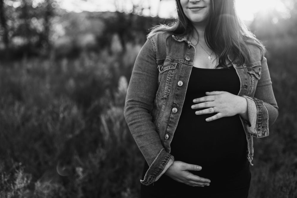 A black and white portrait of an expectant mother wearing a denim jacket while touching her belly outdoors.