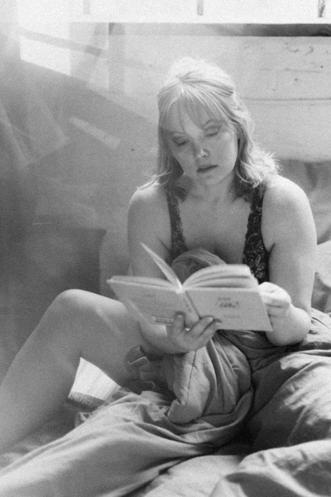 A black and white photo of someone reading a book while relaxing in bed wearing dark loungewear.