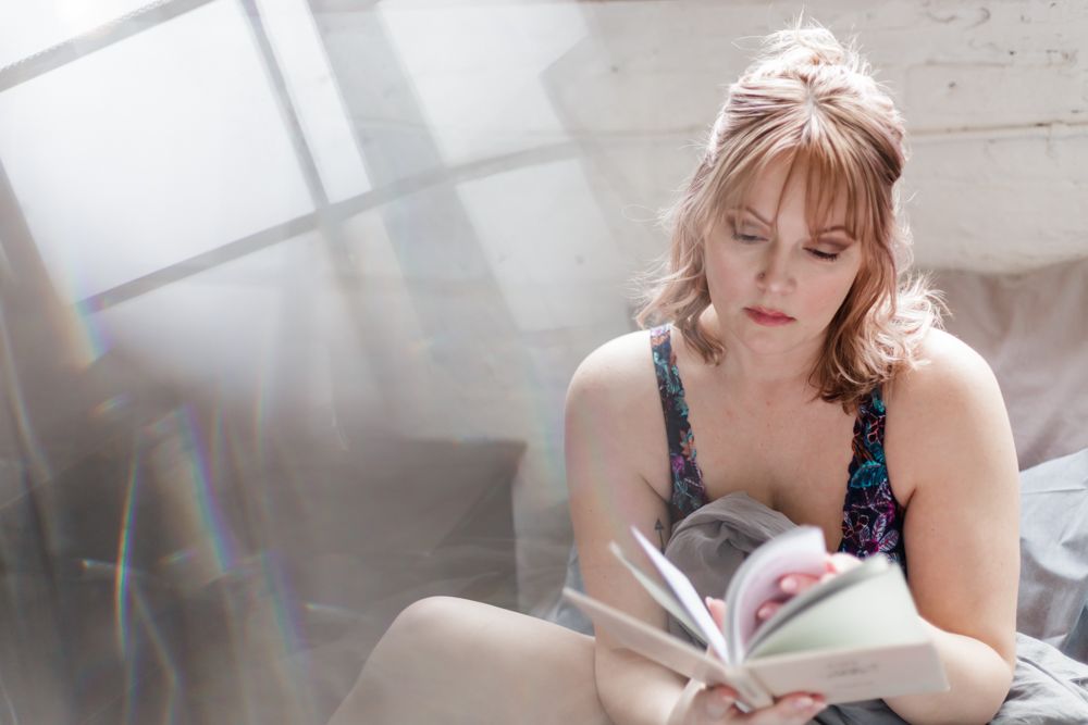Person reading book while relaxing on bed in morning light from window.