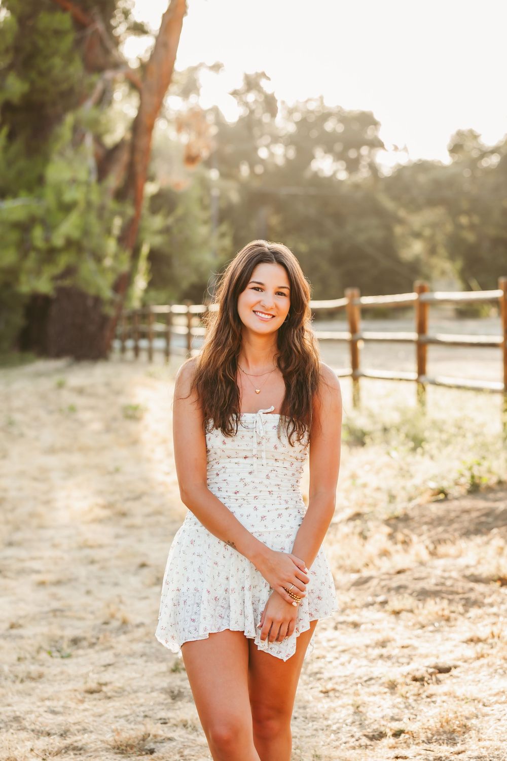 An outdoor portrait session at sunset features a person in a white lace romper posing by a rustic wooden fence.