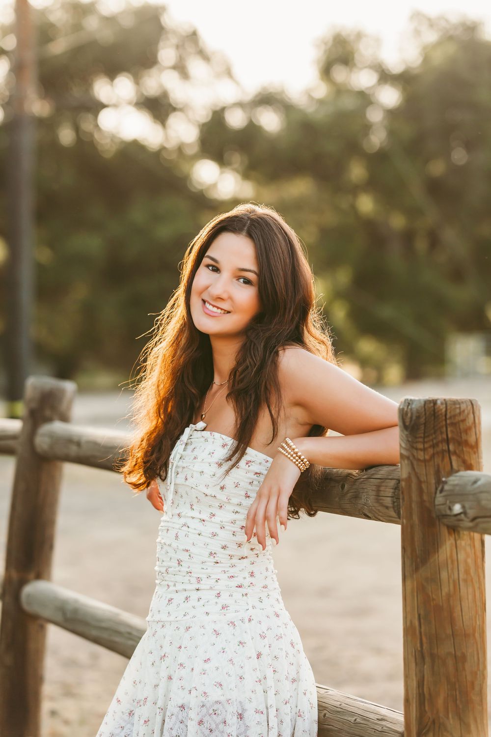 A person in a white floral dress poses along a wooden fence during golden hour sunset in a natural outdoor setting.