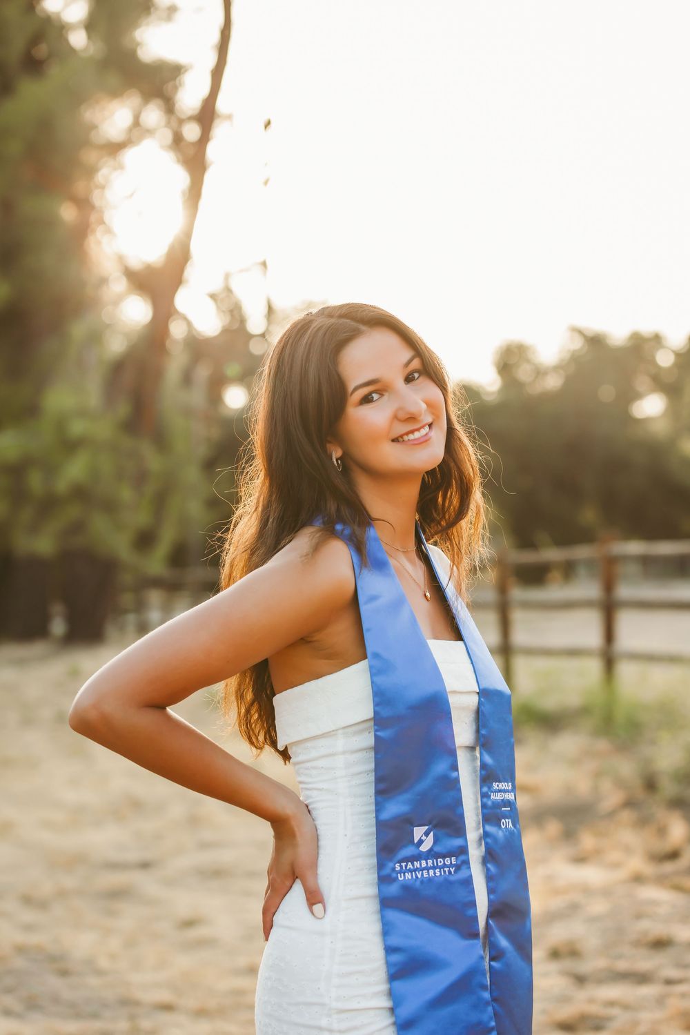 Graduate wearing white dress and blue graduation sash poses outdoors at sunset in natural lighting.