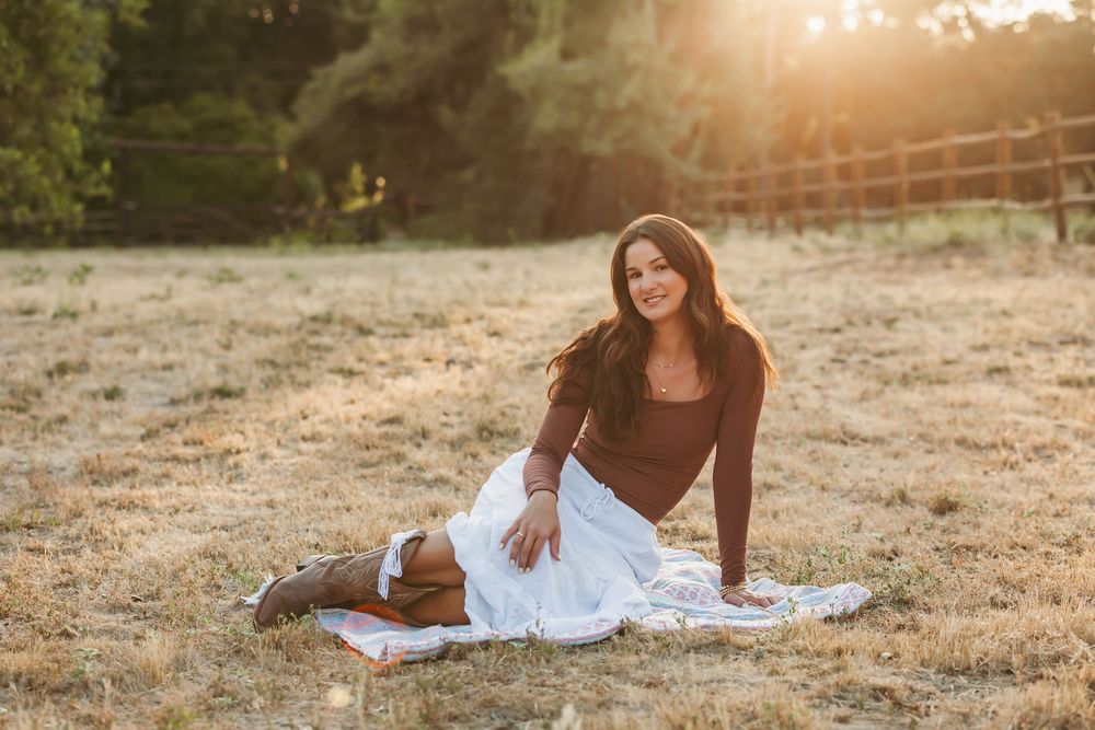 Person relaxing on blanket in sunny field at sunset with coffee thermos and book nearby.
