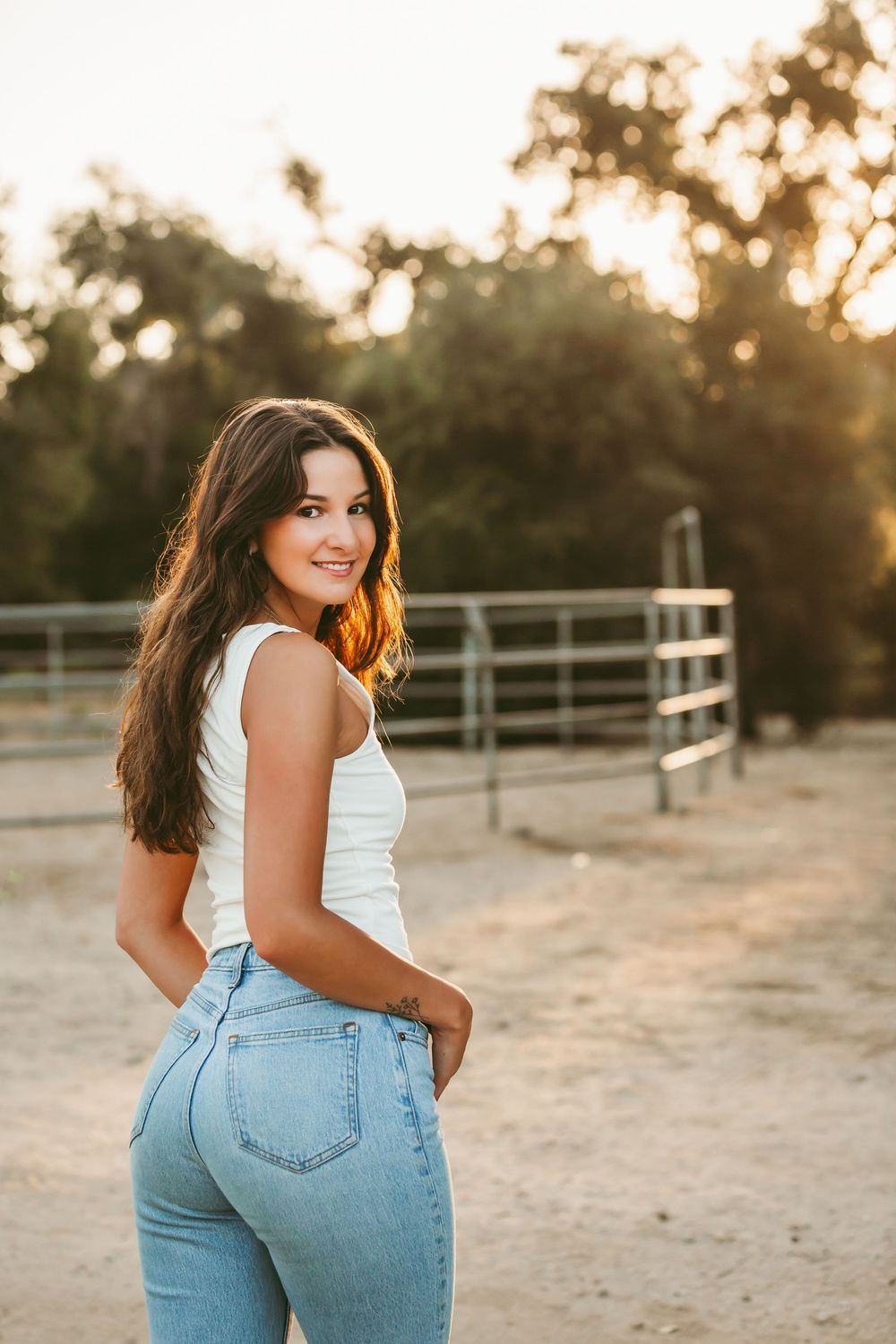 A person in a white tank top and light blue jeans poses at sunset against a fence in a rustic outdoor setting.