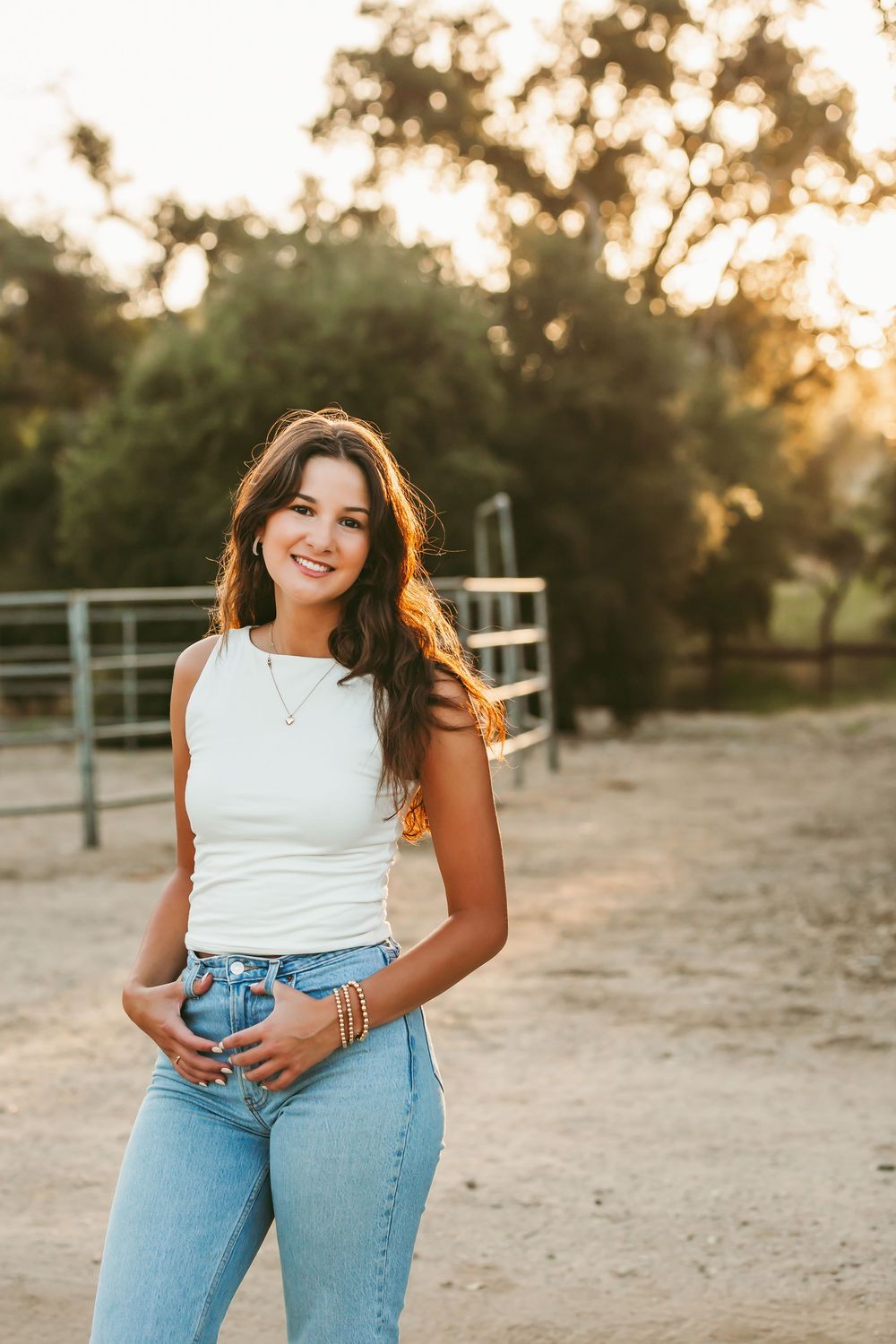 Woman in white top and light blue jeans poses outdoors during golden hour in a rustic setting.