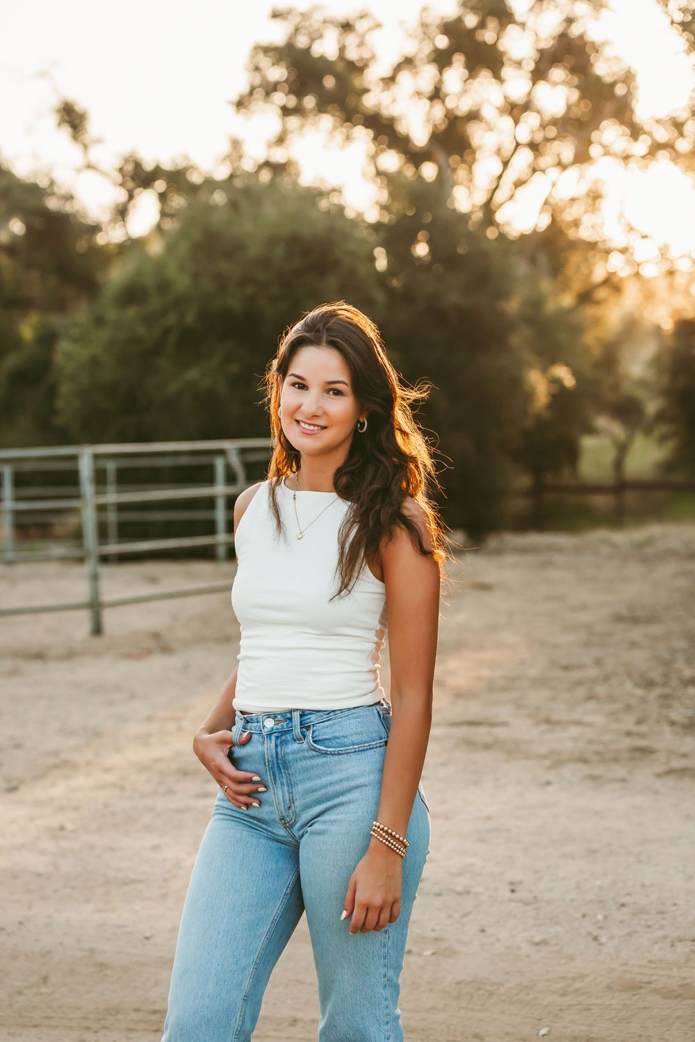 A woman stands in front of a horse pen at golden hour wearing a white tank top and light wash jeans.
