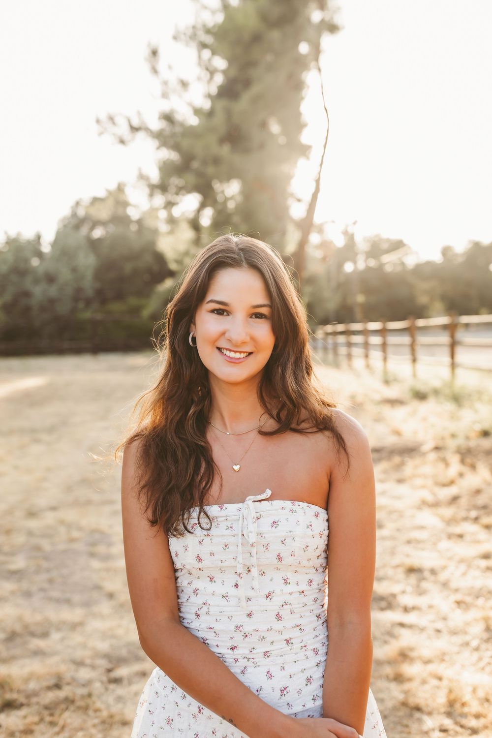 A person in a floral strapless dress smiles at the camera during golden hour with a rustic fence and trees in the background.