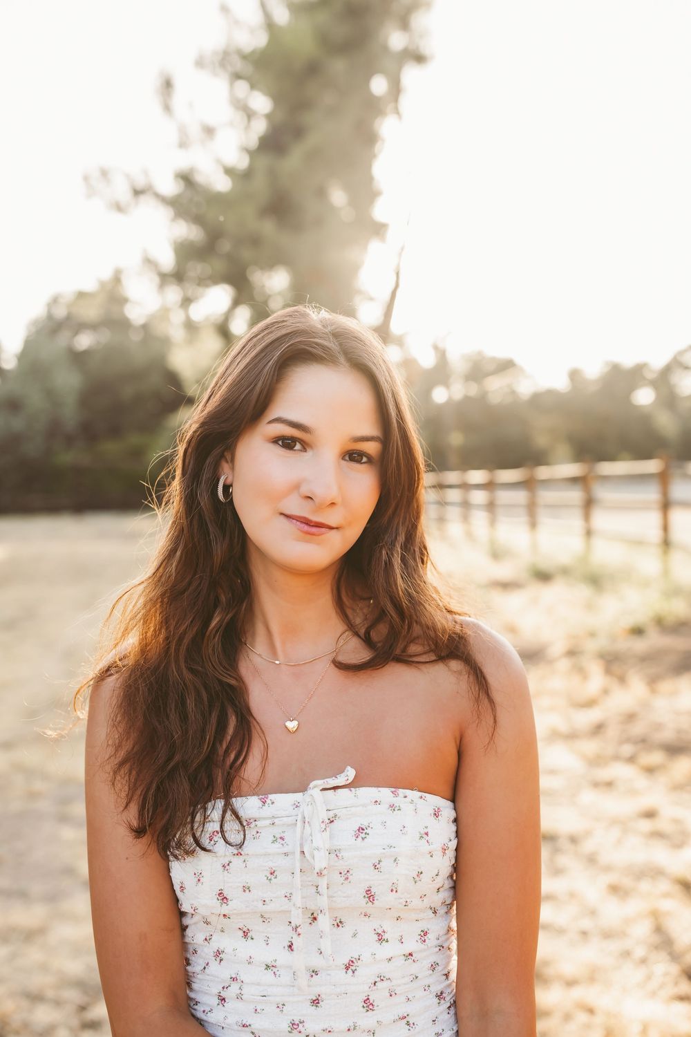 Young woman in white floral dress poses outdoors during golden hour sunset with dreamy backlit lighting effect.