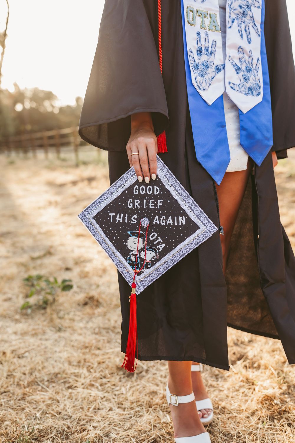 Decorated graduation cap with text 'Good Grief...This Again' shown against black graduation gown and blue stole in sunlight.