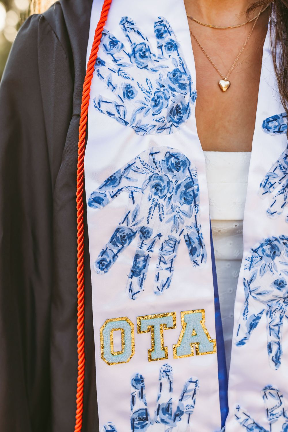 Graduation stole with blue floral handprints and golden 'OTA' text worn with black graduation gown and delicate necklace.