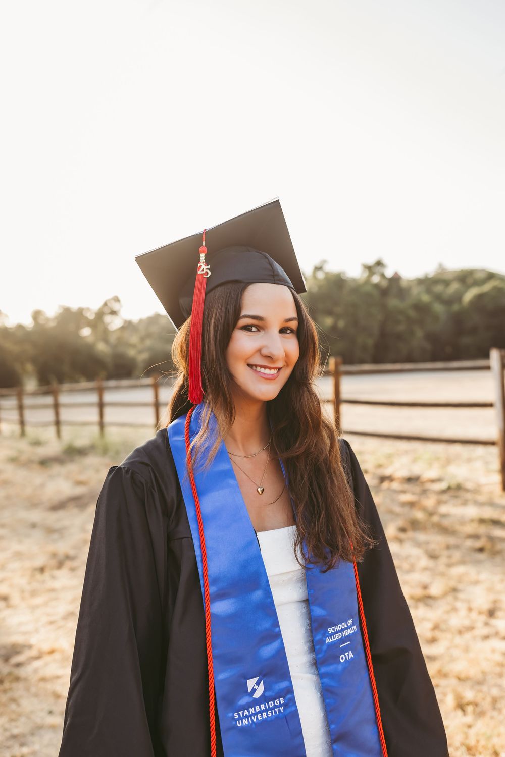 Graduate in cap and gown with blue sash smiling outdoors during golden hour at rustic countryside location.