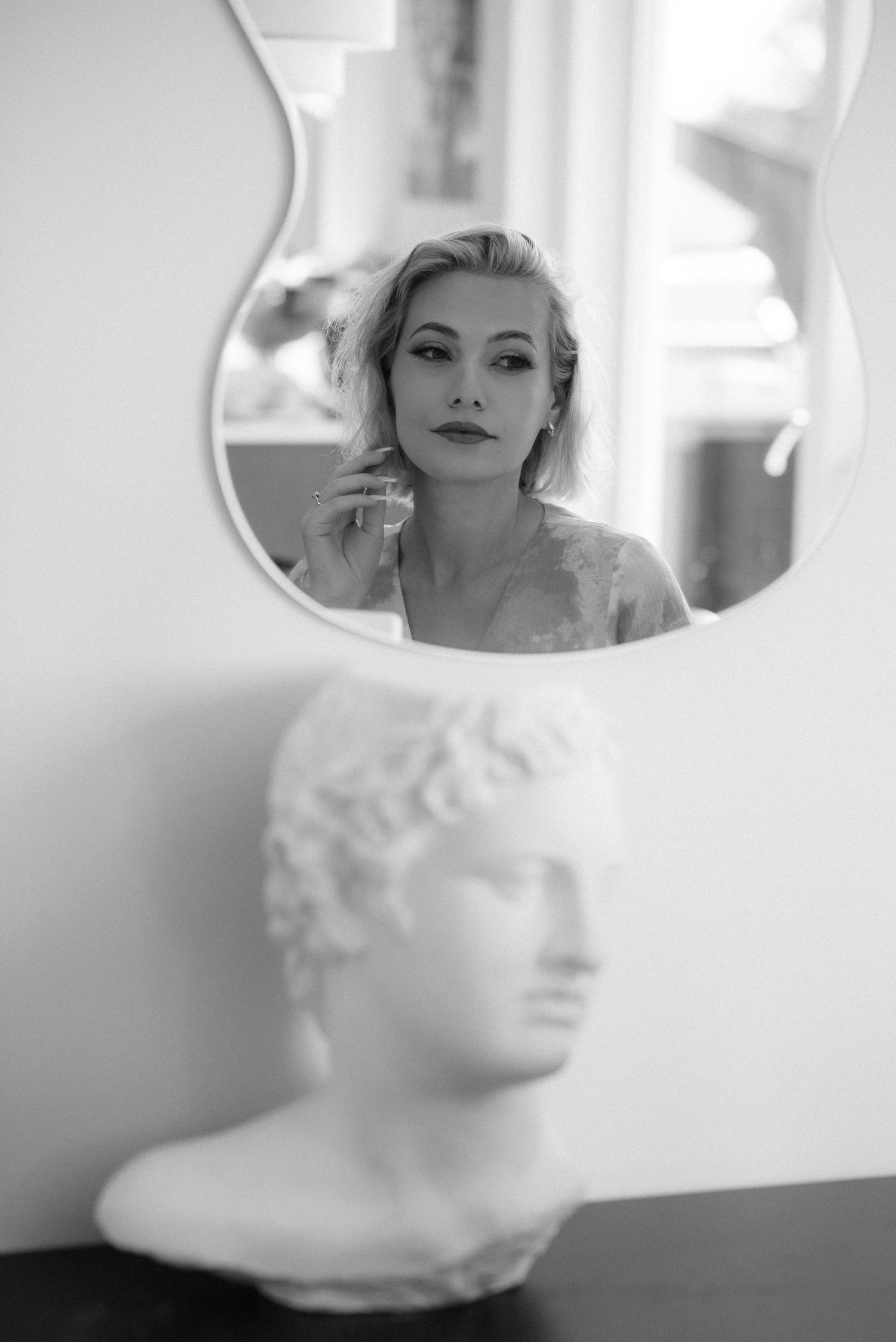 A black and white photograph shows a reflection in a mirror above a classical bust sculpture on a table.