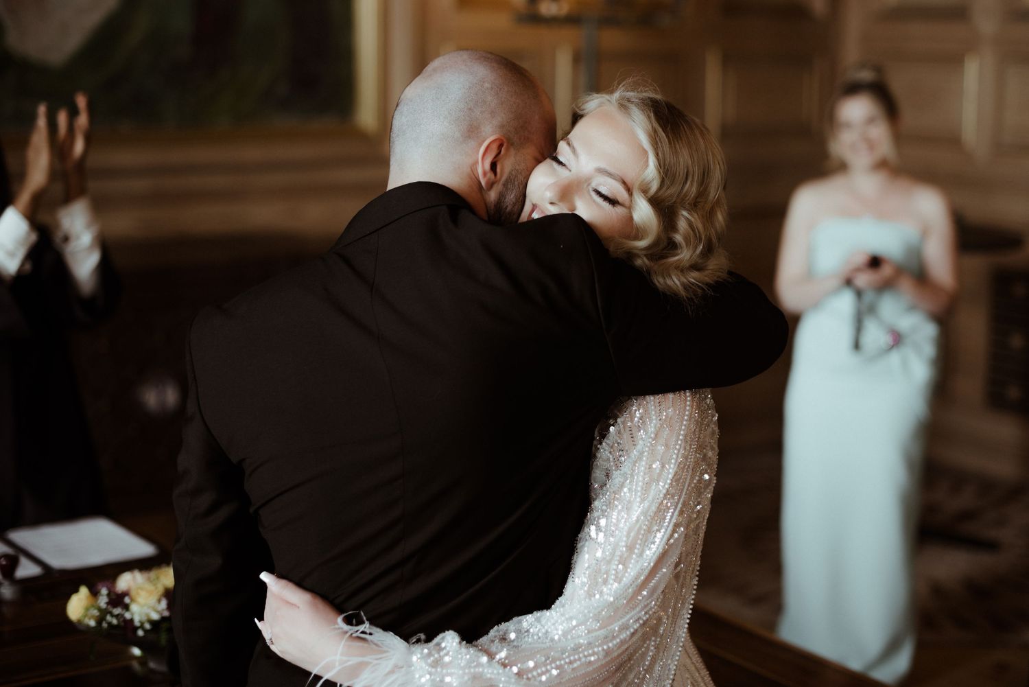 At a formal indoor ceremony, a couple embraces while another attendant in a mint dress stands in the background.
