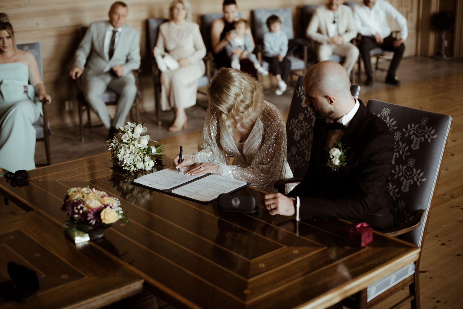 A couple signing legal documents at a wooden table during a wedding ceremony.