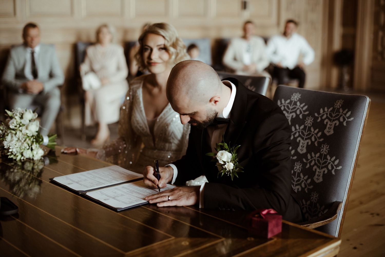 Closeup view of hands signing documents on wooden table with wedding flowers in background.