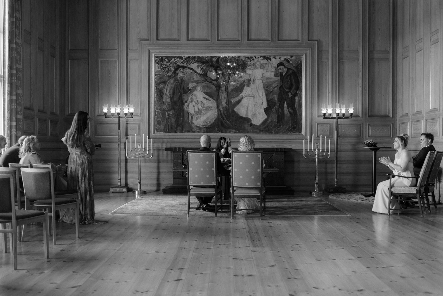 A vintage black and white photograph shows a formal interior room with a large painting and candelabras on display.