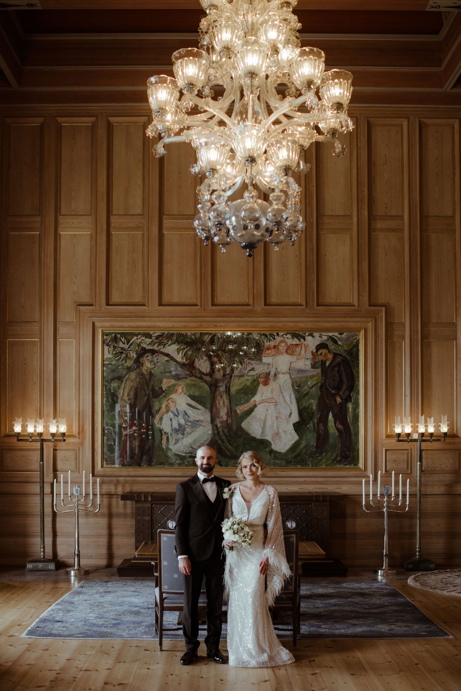 Elegant wedding couple poses beneath crystal chandeliers in ornate ballroom with wood paneling and scenic murals.