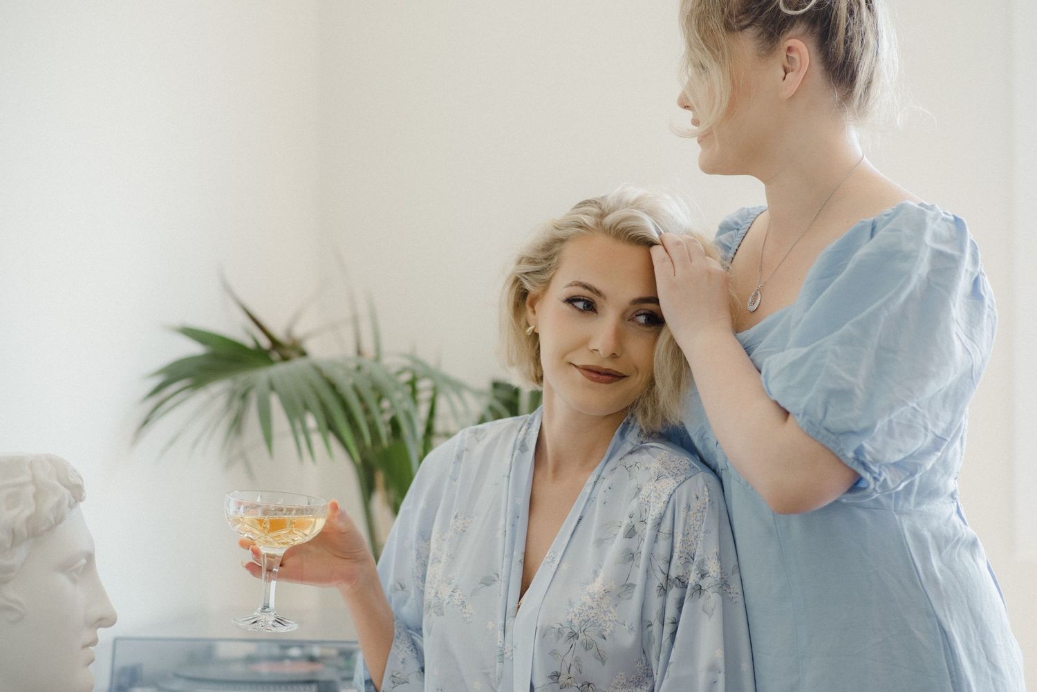Two women in pastel blue robes stand together at home, one holding a cocktail glass near a potted palm plant.