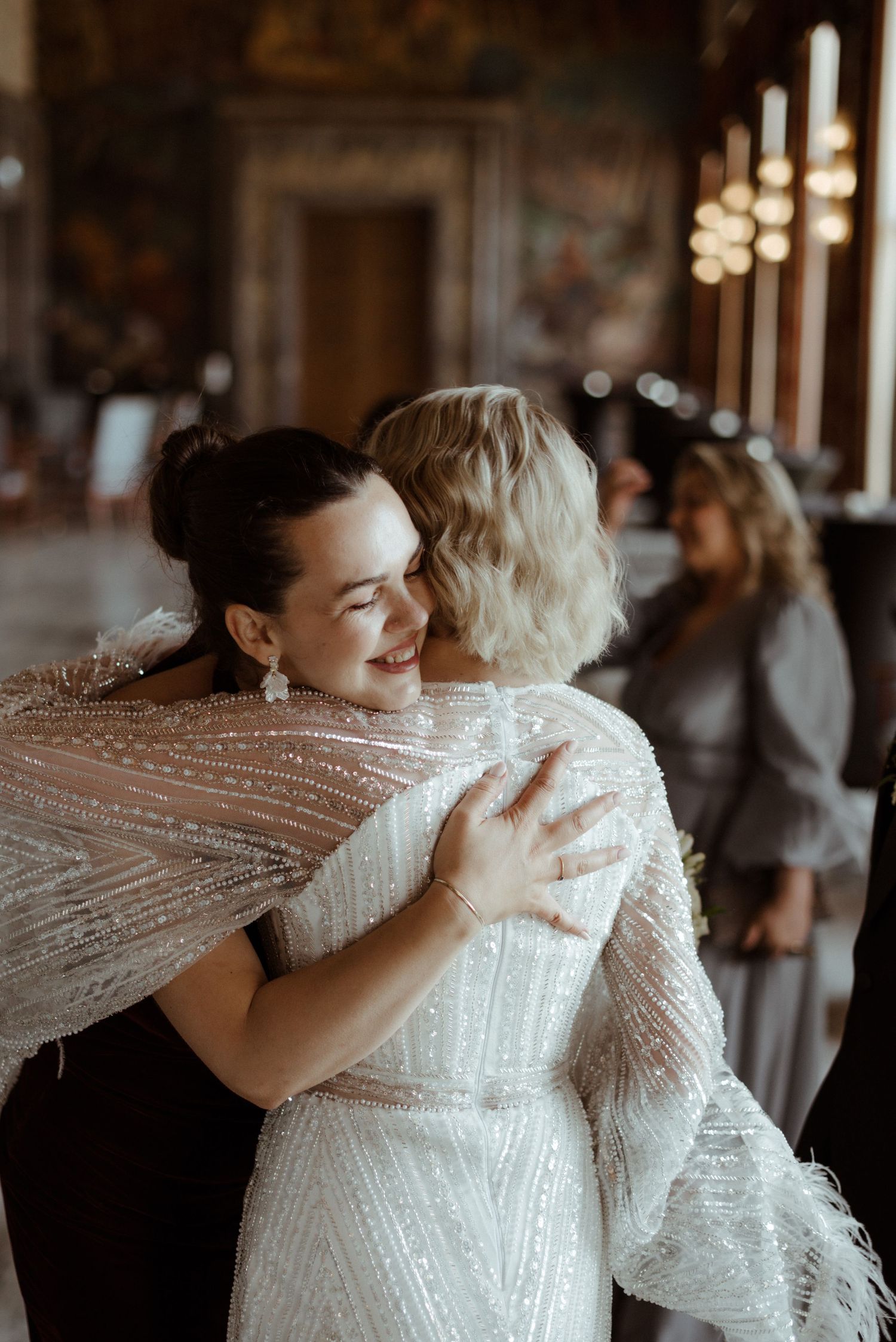 Two people share a joyful embrace at an indoor wedding celebration, wearing elegant white formal attire with sparkles.