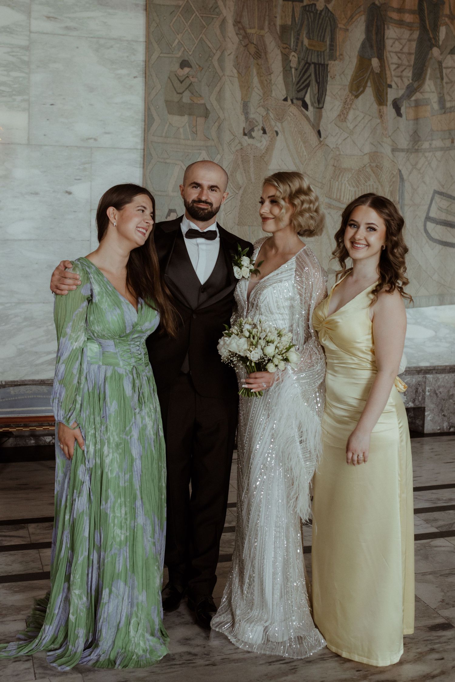 Wedding party poses in elegant attire with the bride in a sparkly white gown and guests in green and yellow dresses.