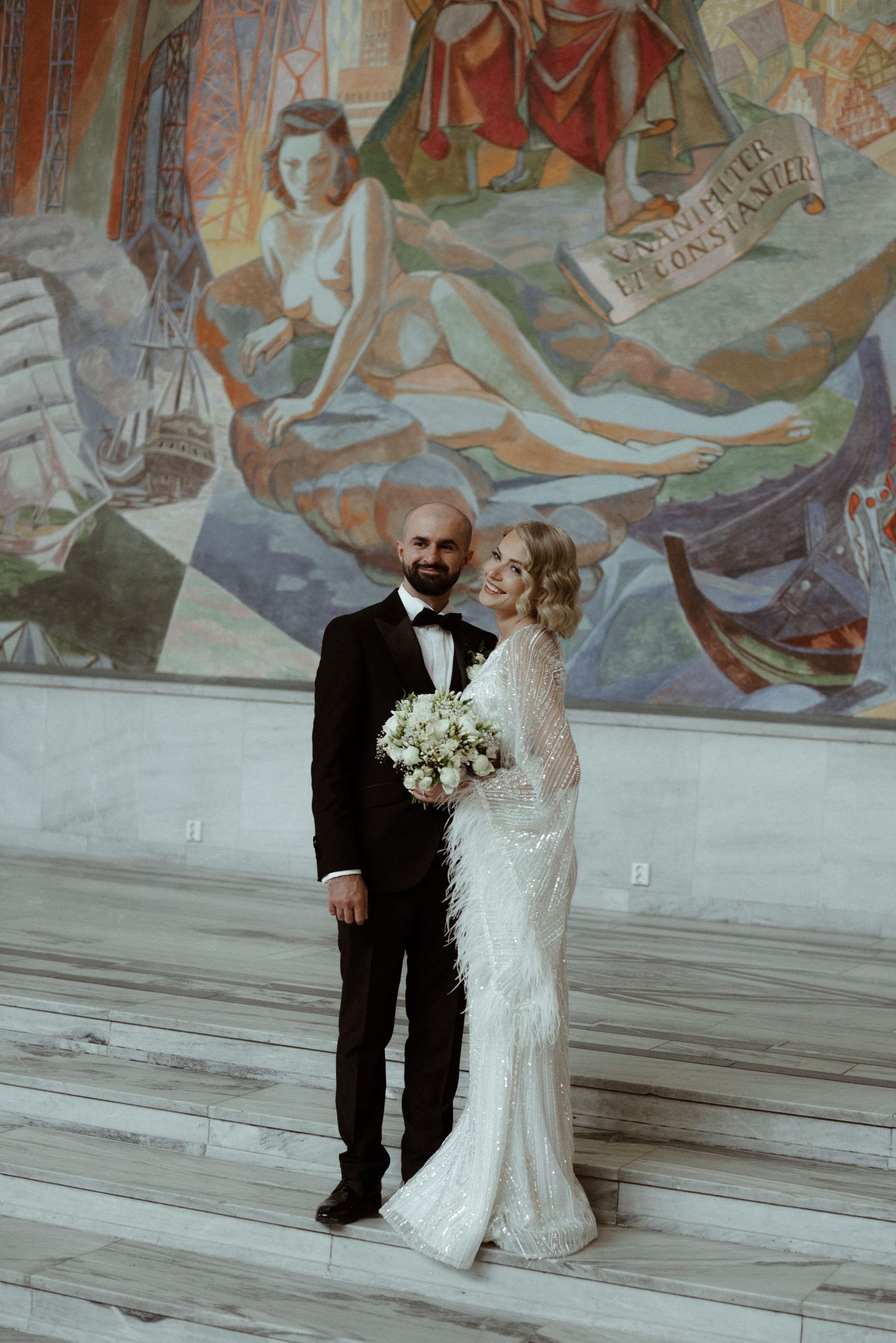A newlywed couple shares a romantic moment against a backdrop of ornate wall murals in a grand historic building.