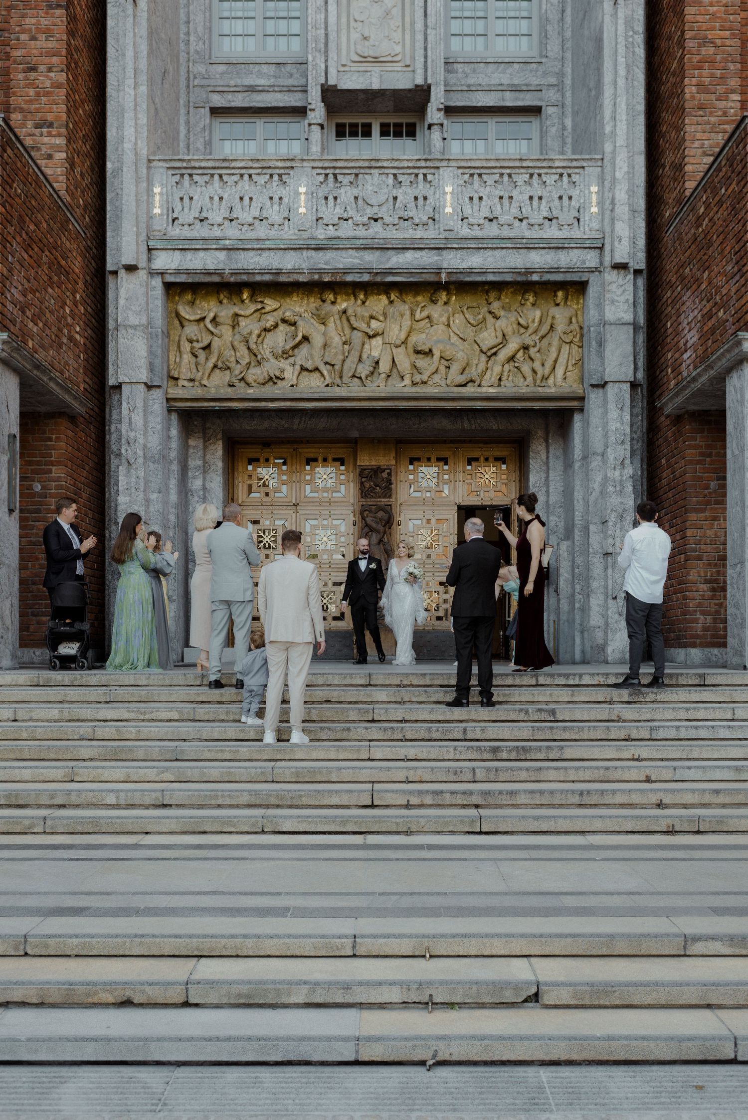 People gather on ornate stone steps of a historic building with brick and carved architectural details.