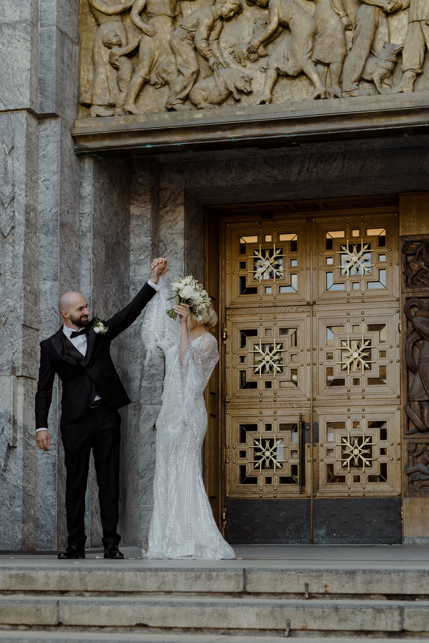 A couple in formal wedding attire celebrate on stone steps in front of an ornate wooden door with decorative carvings.