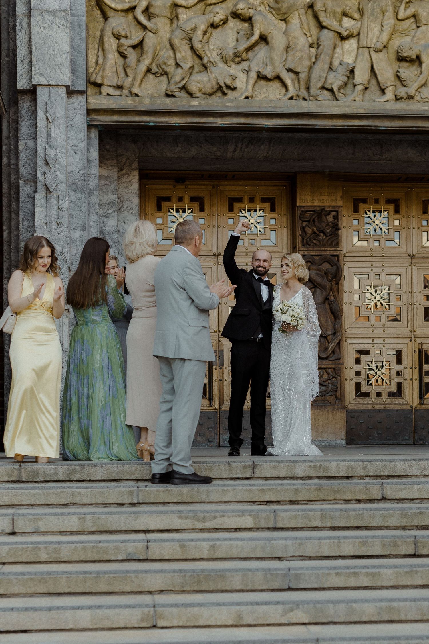 A wedding party stands on stone steps in front of ornate carved doors at a historic building entrance.