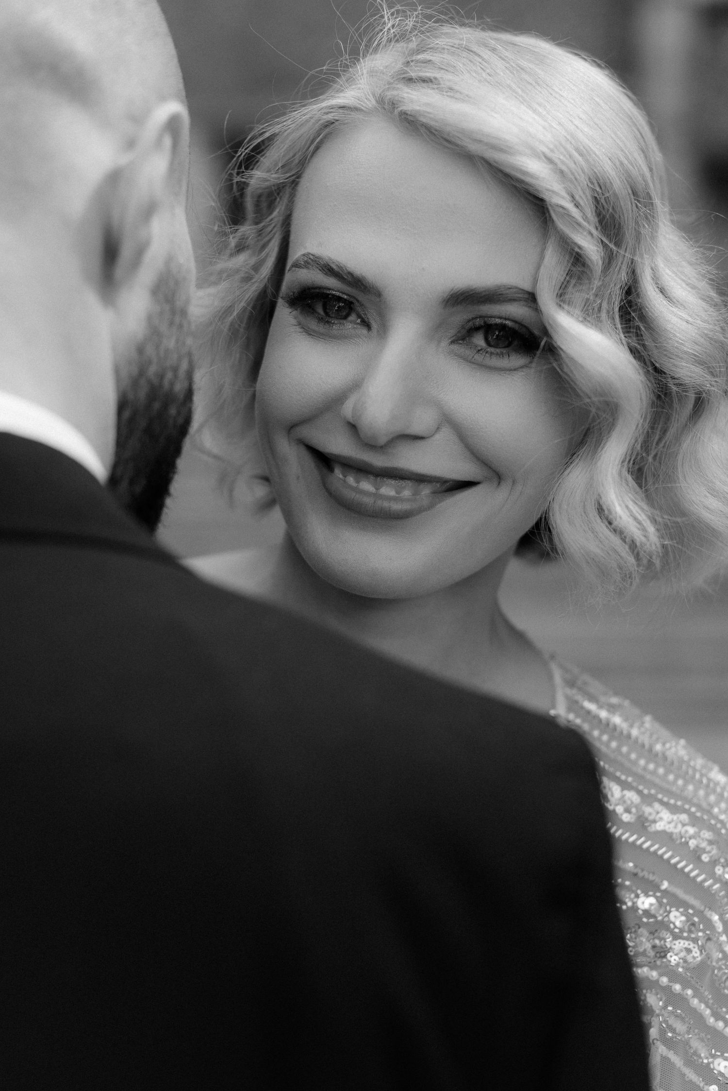 Black and white close-up portrait of a bride with wavy blonde hair smiling while looking at her groom during a wedding moment.