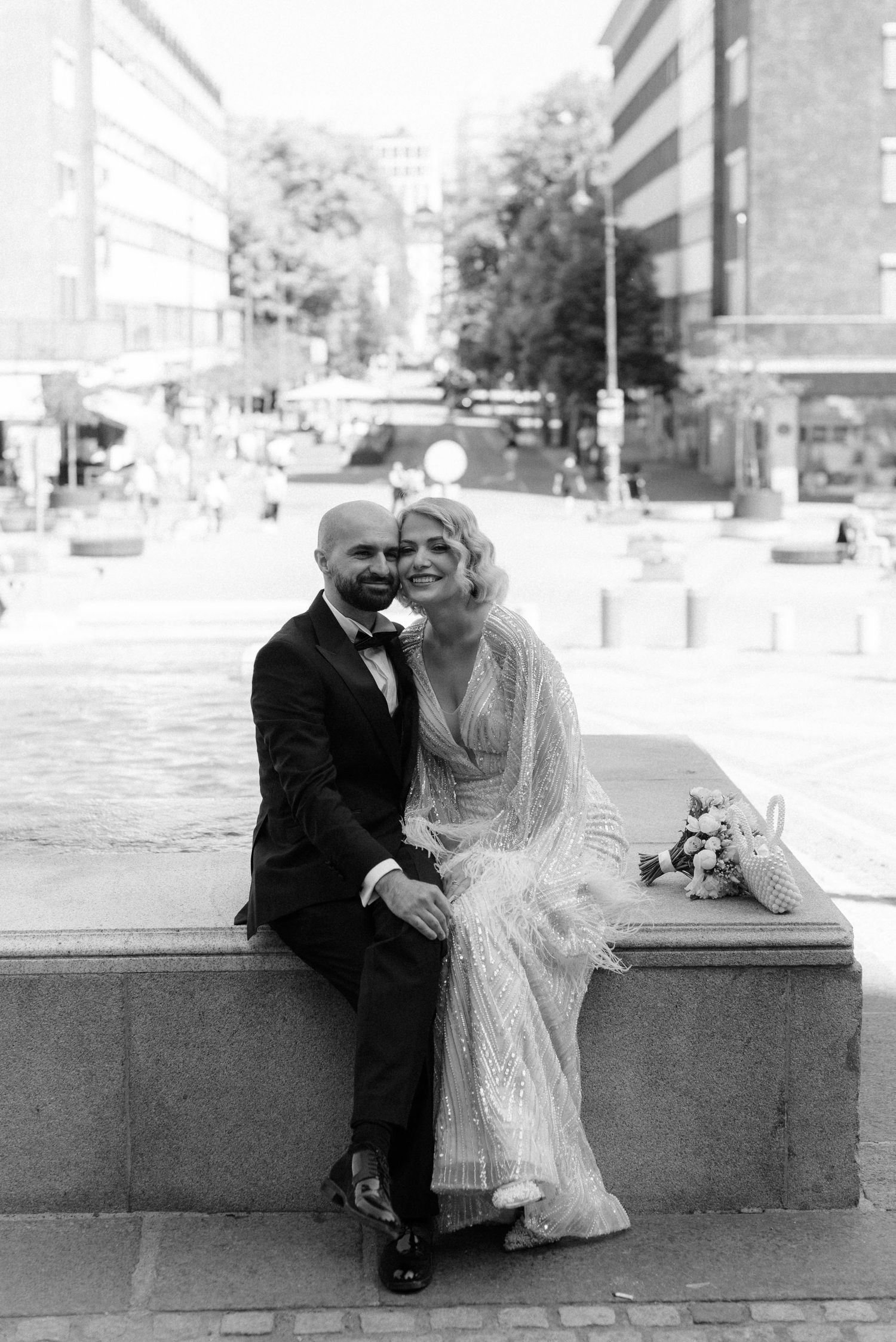 A monochromatic photo of a couple sitting together in wedding attire on an outdoor stone ledge in an urban setting.