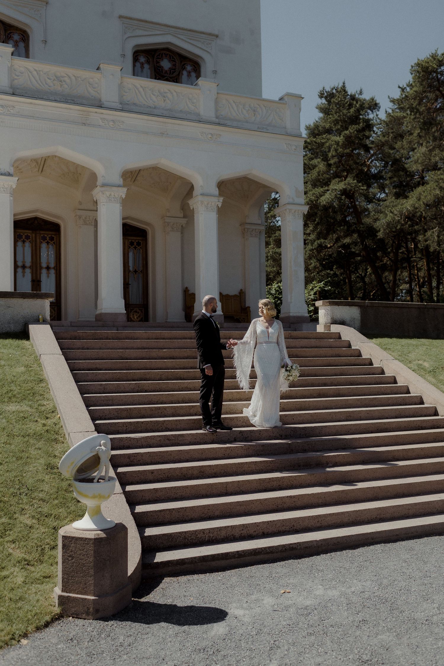 A couple descends the grand stone staircase of a white neoclassical mansion with columns on a sunny day.