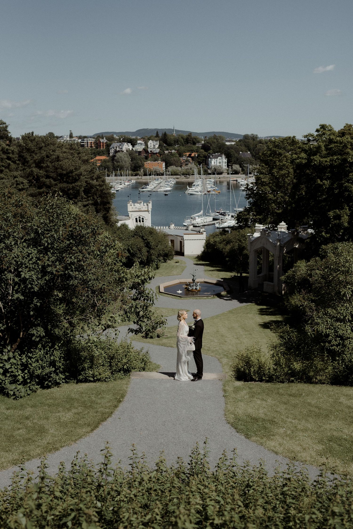 Couples walk along winding paths on a grassy hillside overlooking a scenic harbor with boats and coastal buildings.
