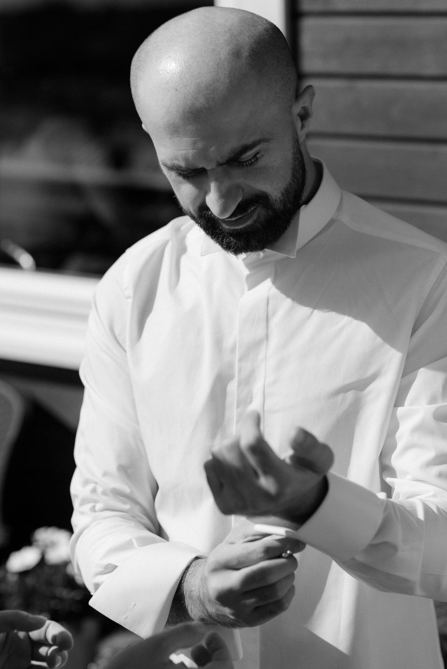 A black and white photo of someone in a white dress shirt buttoning their cuff while smiling downward.