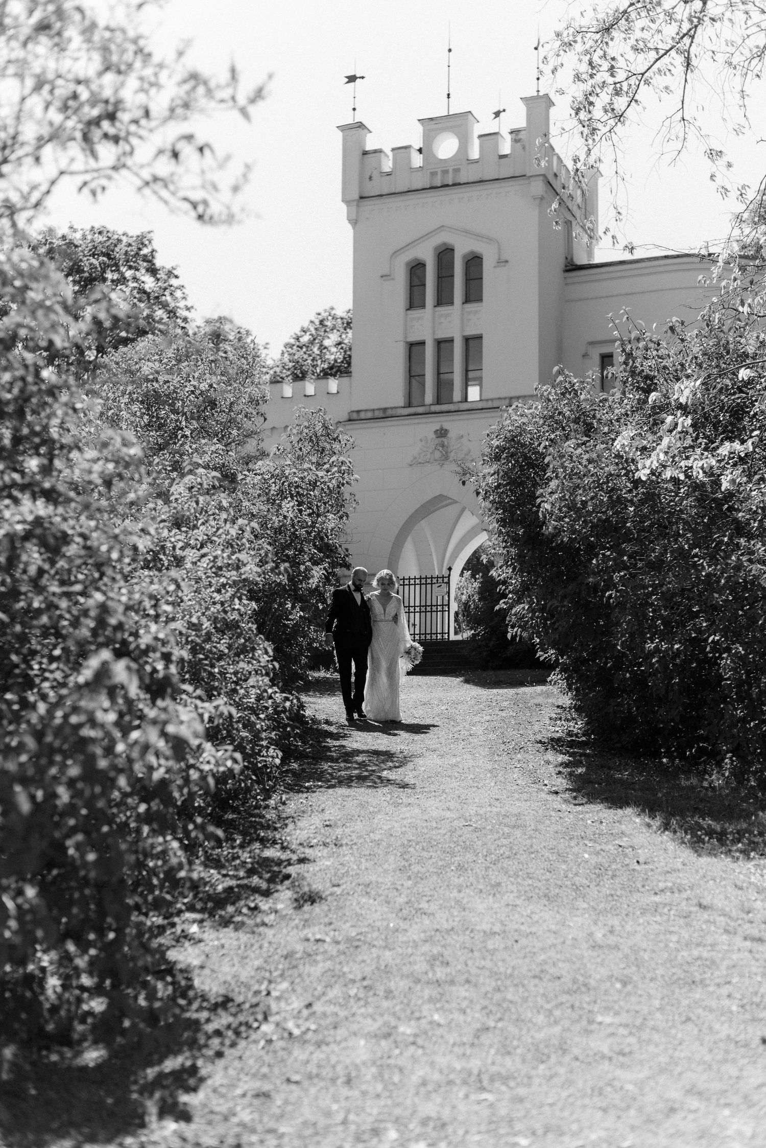 A black and white photograph showing a castle-like building with arches at the end of a garden path lined with shrubs.