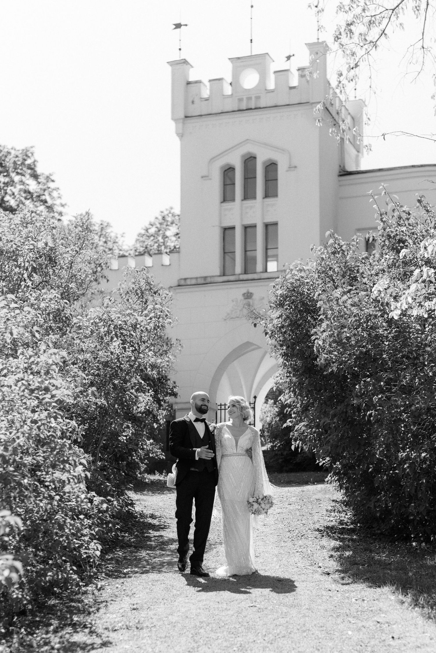 A couple walks together along a garden path in front of a white castle-like building with Gothic architecture.