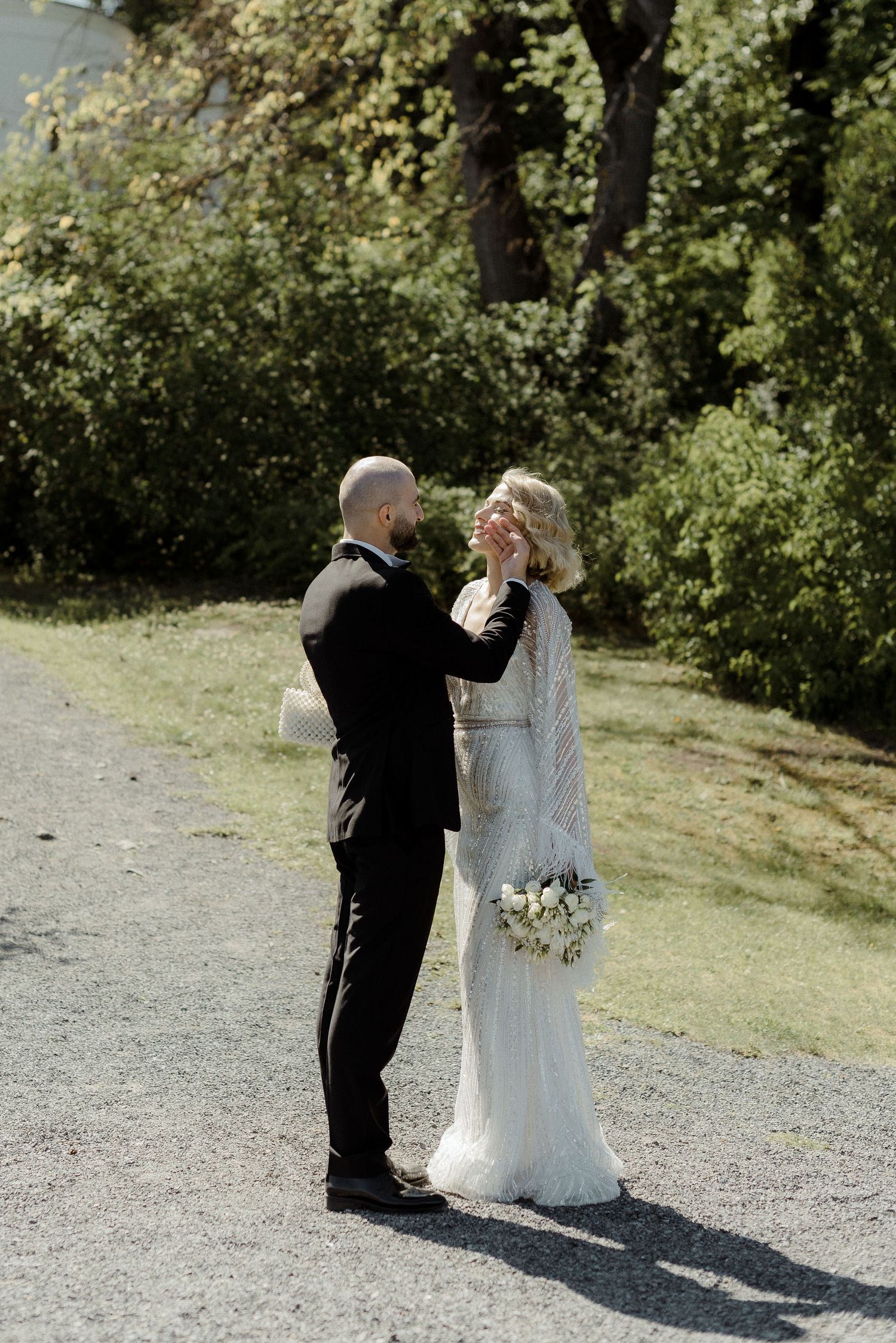 A wedding couple shares an intimate moment on a garden path, with the bride in white lace and groom in a dark suit.