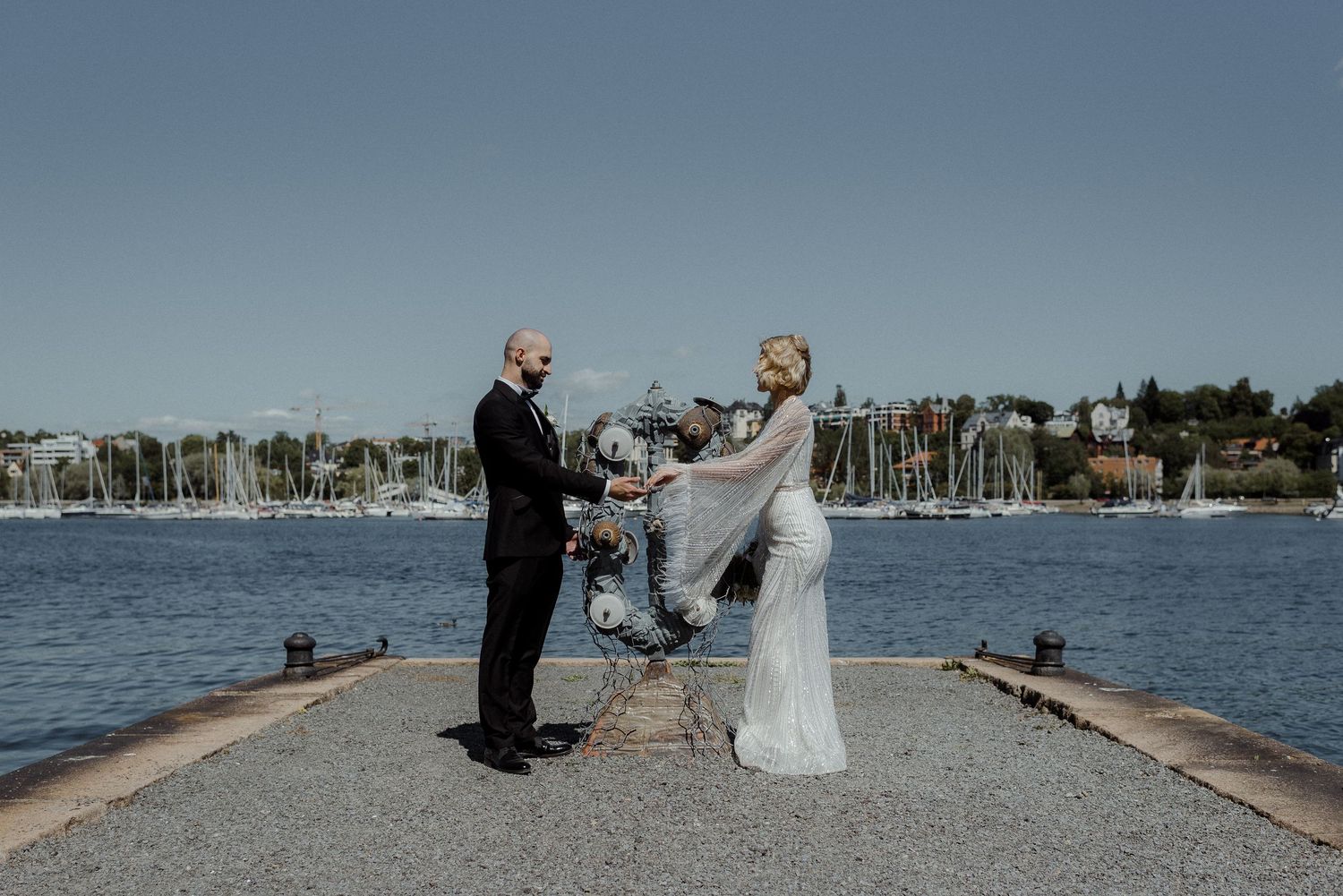 A small wedding ceremony taking place on a coastal pier overlooking a harbor on a sunny afternoon.