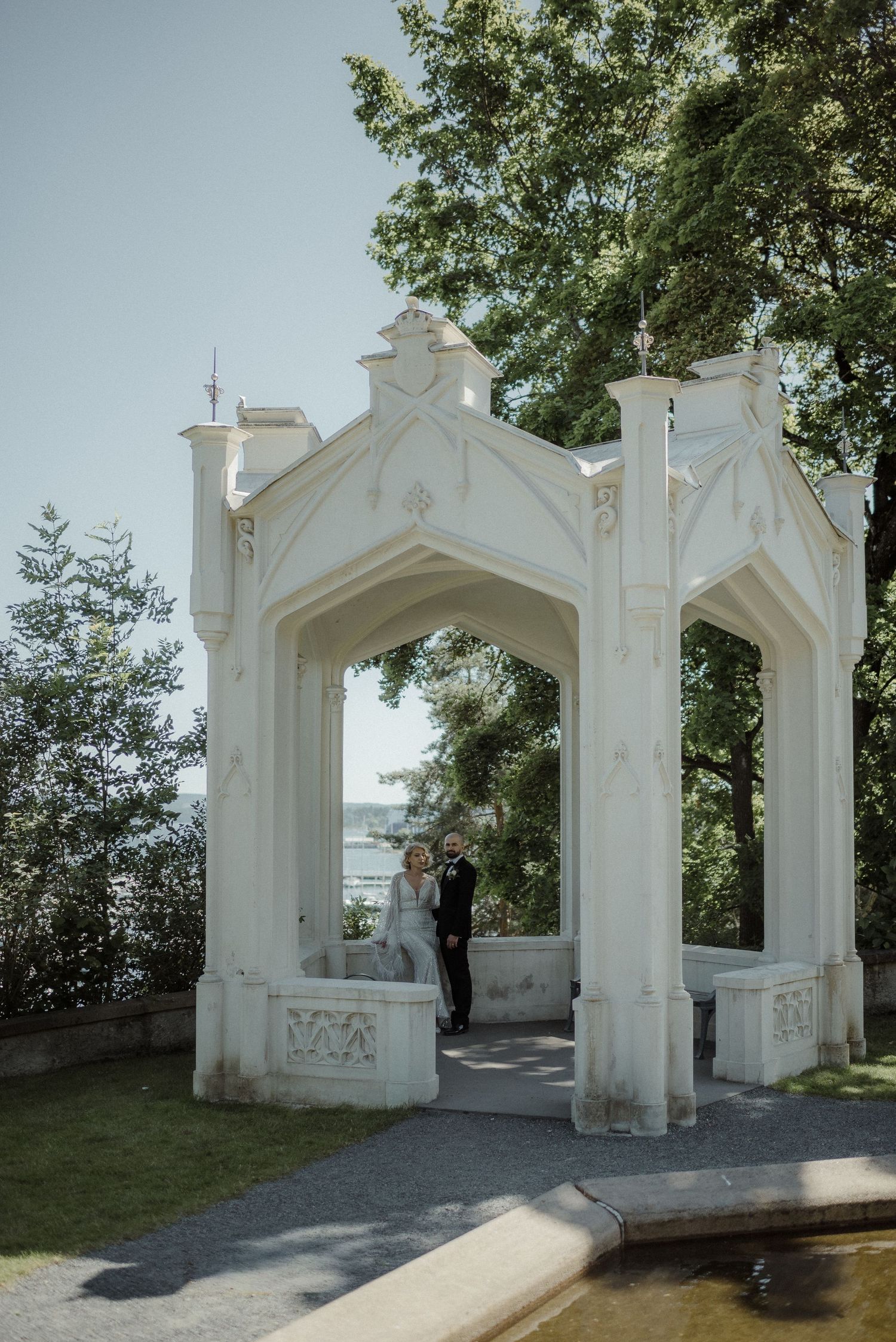A beautiful white Gothic-style gazebo with ornate architectural details stands in a garden overlooking the water.